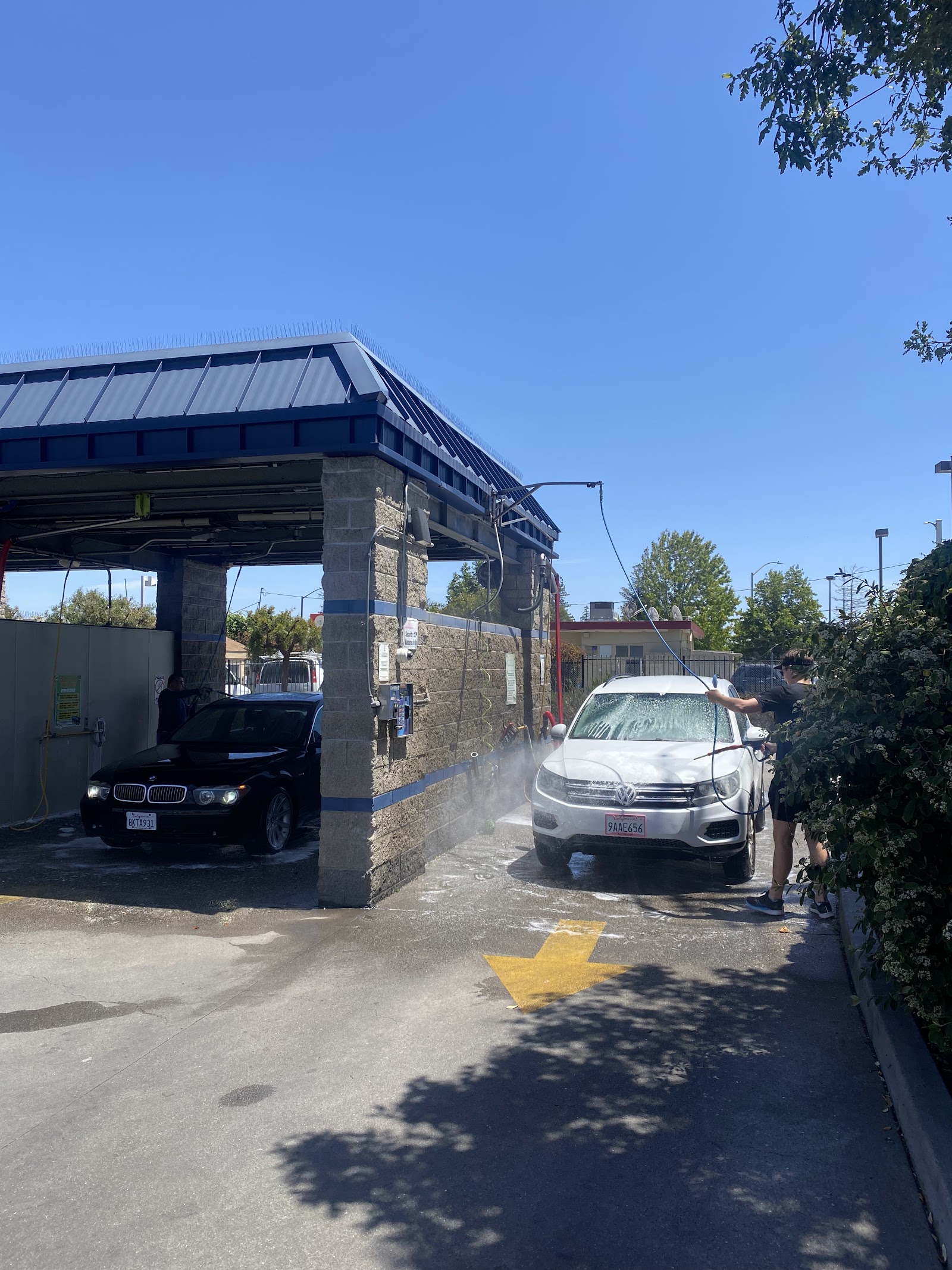 A Clear Water Car Wash bay with a black BMW and a white VW being washed under a stone-and-blue-trim canopy on a sunny Peninsula day